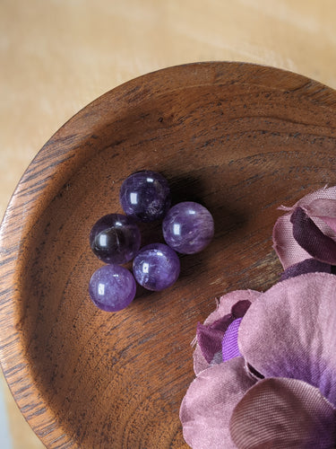 Tiny amethyst spheres in a wooden bowl next to purple silk flowers.