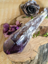 Load image into Gallery viewer, Large amethyst wand laying across two wooden stumps with periwinkle leaves and black and purple silk flowers
