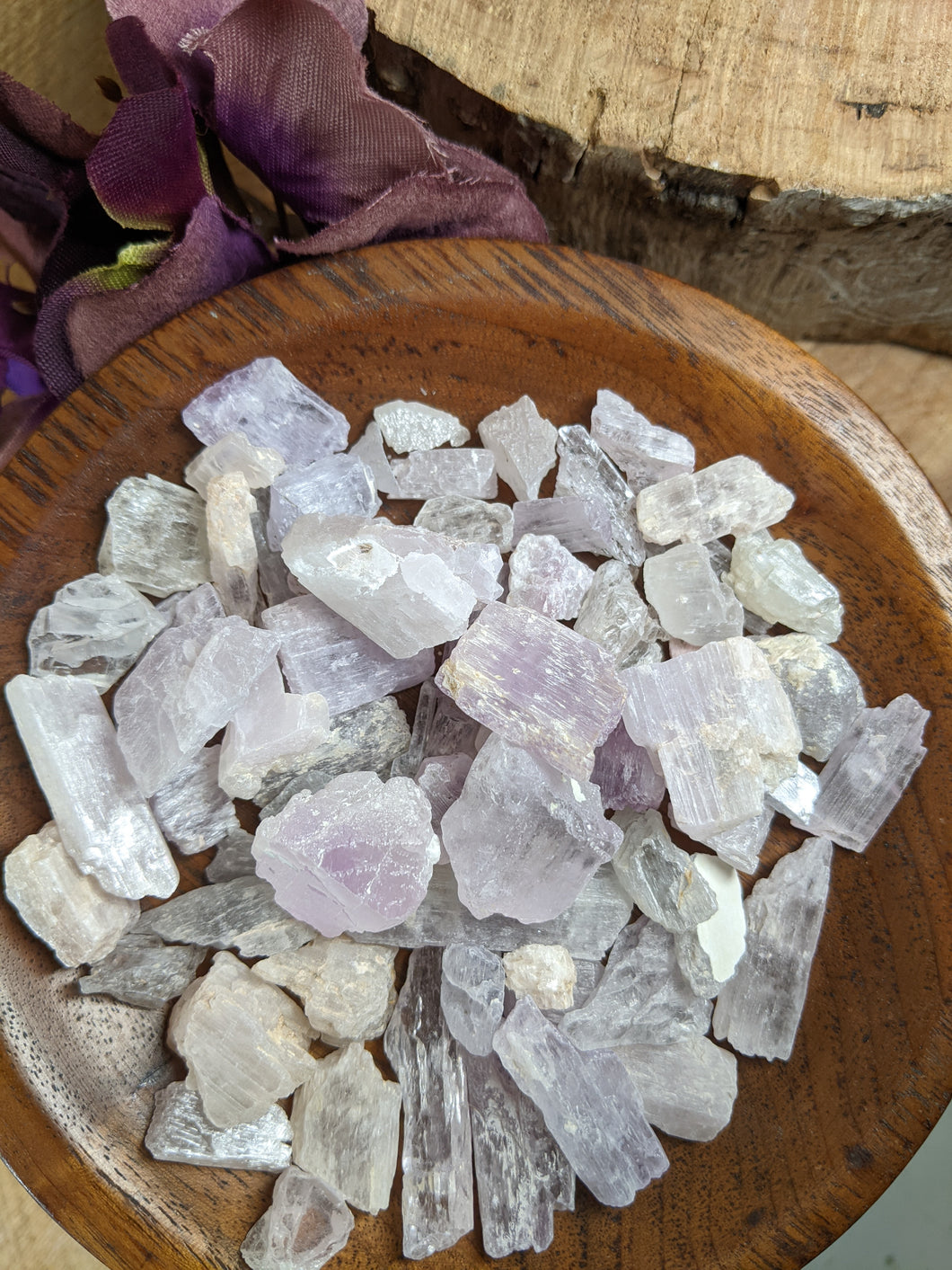 Mixed pieces of raw kunzite in a wooden bowl, next to a purple silk flower and part of a tree stump.